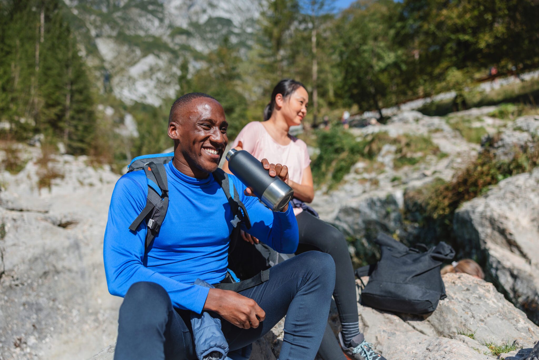 Two friends, a Black man and an Asian woman, rest and hydrate while hiking in a rocky mountain area on a sunny day.
